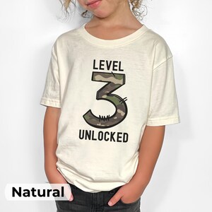 May include: A cream-colored t-shirt with the words "LEVEL 3 UNLOCKED" printed in black. The number 3 is filled with a camouflage pattern. The shirt is worn by a child.