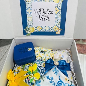 Peut inclure: Un coffret cadeau avec un motif floral bleu et blanc, contenant divers articles. La boîte comprend une boîte à bijoux bleue, un chouchou, une écharpe et un nœud. La boîte porte l'inscription "La Dolce Vita" sur le couvercle.