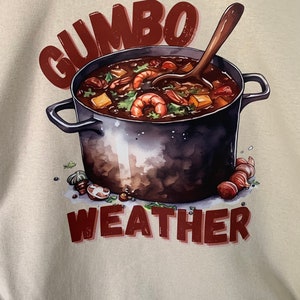 May include: A beige t-shirt with a brown illustration of a pot of gumbo with a wooden spoon. The text "GUMBO WEATHER" is printed in brown below the pot.