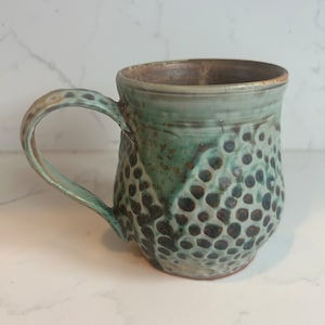 Puede incluir: Una taza de cerámica hecha a mano con un esmalte verde moteado y un asa marrón. La taza tiene un diseño texturizado único con puntos en relieve.