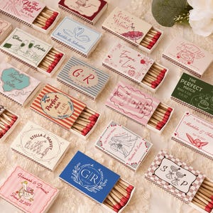 Puede incluir: Una colección de cajas de cerillas rectangulares con varios diseños impresos personalizados, que incluyen nombres, fechas e ilustraciones como cisnes, copas de champán y motivos florales. Cada caja está parcialmente abierta, mostrando cerillas de madera.