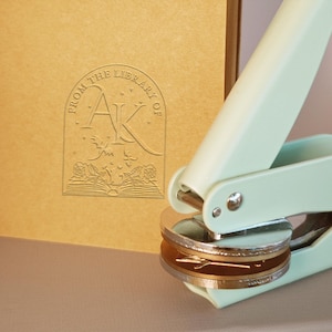 Può includere: Un'incisoria per libri verde menta accanto a un libro marrone con il testo "FROM THE LIBRARY OF AK" in un design decorativo. L'incisoria ha una matrice argentata e dorata.