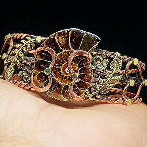 K&ouml;nnte beinhalten: Ein handgefertigtes Kupferarmband mit einem Ammoniten-Fossil-Mittelst&uuml;ck. Das Armband weist aufwendige Blumen- und Blattdesigns in Bronze- und Goldt&ouml;nen mit einem gedrehten Kupferband auf. Das Fossil hat eine satte braune Farbe mit detaillierten Mustern.