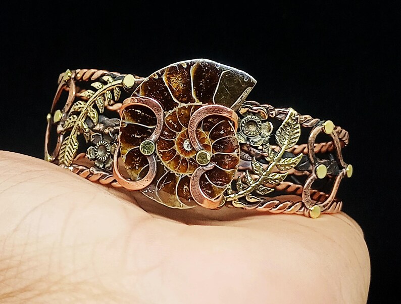 K&ouml;nnte beinhalten: Ein handgefertigtes Kupferarmband mit einem Ammoniten-Fossil-Mittelst&uuml;ck. Das Armband weist aufwendige Blumen- und Blattdesigns in Bronze- und Goldt&ouml;nen mit einem gedrehten Kupferband auf. Das Fossil hat eine satte braune Farbe mit detaillierten Mustern.
