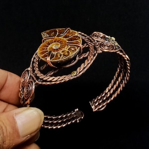 May include: A copper wire-wrapped cuff bracelet featuring a central brown ammonite fossil. The metalwork consists of twisted wire strands, circular coil accents, and a secure setting holding the fossil in place, creating an open-ended design.