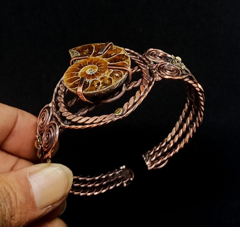 May include: A copper wire-wrapped cuff bracelet featuring a central brown ammonite fossil. The metalwork consists of twisted wire strands, circular coil accents, and a secure setting holding the fossil in place, creating an open-ended design.