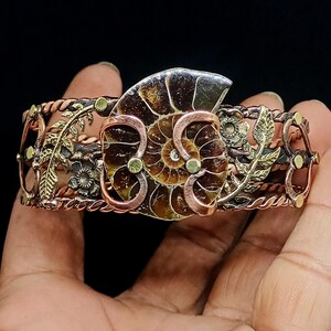 K&ouml;nnte beinhalten: Handgefertigtes Kupfer- und Messing-Armreif-Armband mit einem Ammoniten-Fossil-Mittelst&uuml;ck. Das Armband hat aufwendige Blumen- und Blattmuster mit einer Mischung aus Kupfer-, Messing- und Goldt&ouml;nen. Das Fossil ist hellbraun mit einem Spiralmuster.