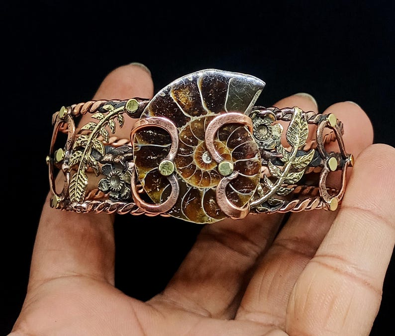 K&ouml;nnte beinhalten: Handgefertigtes Kupfer- und Messing-Armreif-Armband mit einem Ammoniten-Fossil-Mittelst&uuml;ck. Das Armband hat aufwendige Blumen- und Blattmuster mit einer Mischung aus Kupfer-, Messing- und Goldt&ouml;nen. Das Fossil ist hellbraun mit einem Spiralmuster.