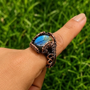 May include: A wire-wrapped ring featuring a central oval labradorite gemstone with a blue iridescent flash. The setting is crafted from dark copper wire, intricately coiled and looped around the stone to create a textured, metallic band.