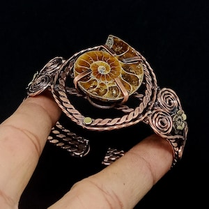 May include: A copper wire cuff bracelet featuring a central spiral ammonite fossil. The metalwork includes twisted wire bands, circular scroll patterns, and small metallic accents. The bracelet has an open design with a dark, oxidised finish.