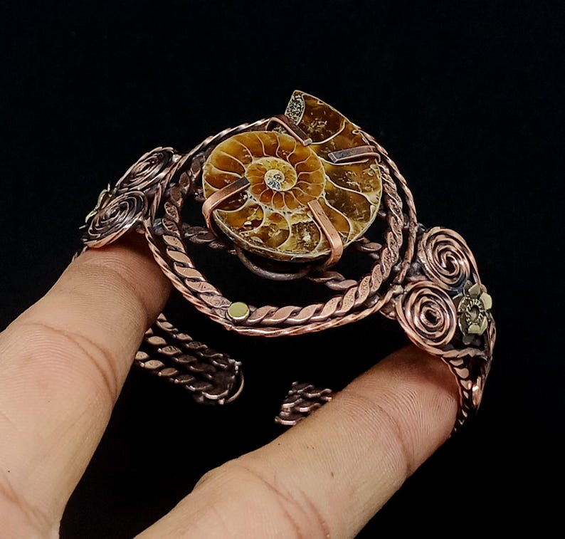 May include: A copper wire cuff bracelet featuring a central spiral ammonite fossil. The metalwork includes twisted wire bands, circular scroll patterns, and small metallic accents. The bracelet has an open design with a dark, oxidised finish.