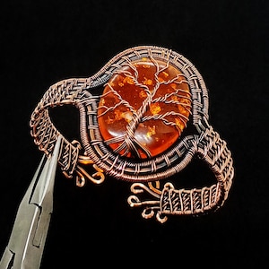 Puede incluir: Un brazalete artesanal de alambre de cobre con una piedra de ámbar ovalada con un diseño de árbol. El brazalete tiene un intrincado trabajo de alambre y una paleta de colores cálidos y terrosos. La piedra de ámbar mide aproximadamente 3,8 cm de largo.