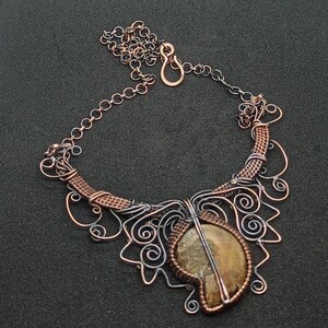 K&ouml;nnte beinhalten: Eine Halskette aus Kupfer- und Silberdraht mit einem Anh&auml;nger aus braunem Achat. Die Halskette hat ein zartes, filigranes Design mit Spiralen und Schleifen.