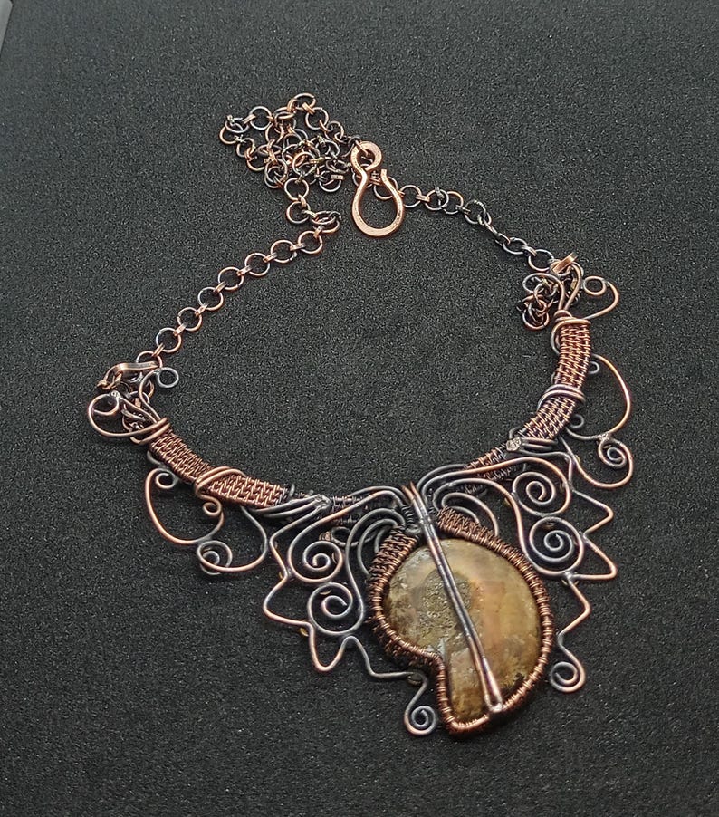 K&ouml;nnte beinhalten: Eine Halskette aus Kupfer- und Silberdraht mit einem Anh&auml;nger aus braunem Achat. Die Halskette hat ein zartes, filigranes Design mit Spiralen und Schleifen.