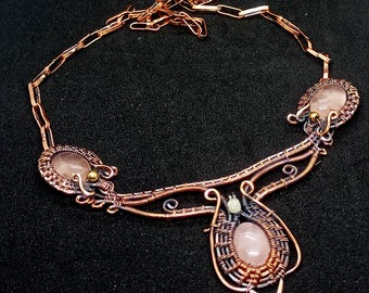 May include: A handcrafted copper wire necklace featuring three oval rose quartz stones. The necklace has a woven wire design around the stones and a chain closure. The necklace is displayed on a black surface.