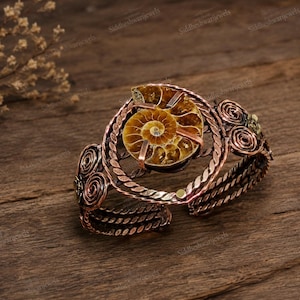 May include: A copper-toned cuff bracelet featuring a central spiral-patterned ammonite fossil held by metal prongs. The band is constructed from twisted wire and decorated with symmetrical wire-wrapped swirls on both sides of the fossil centrepiece.