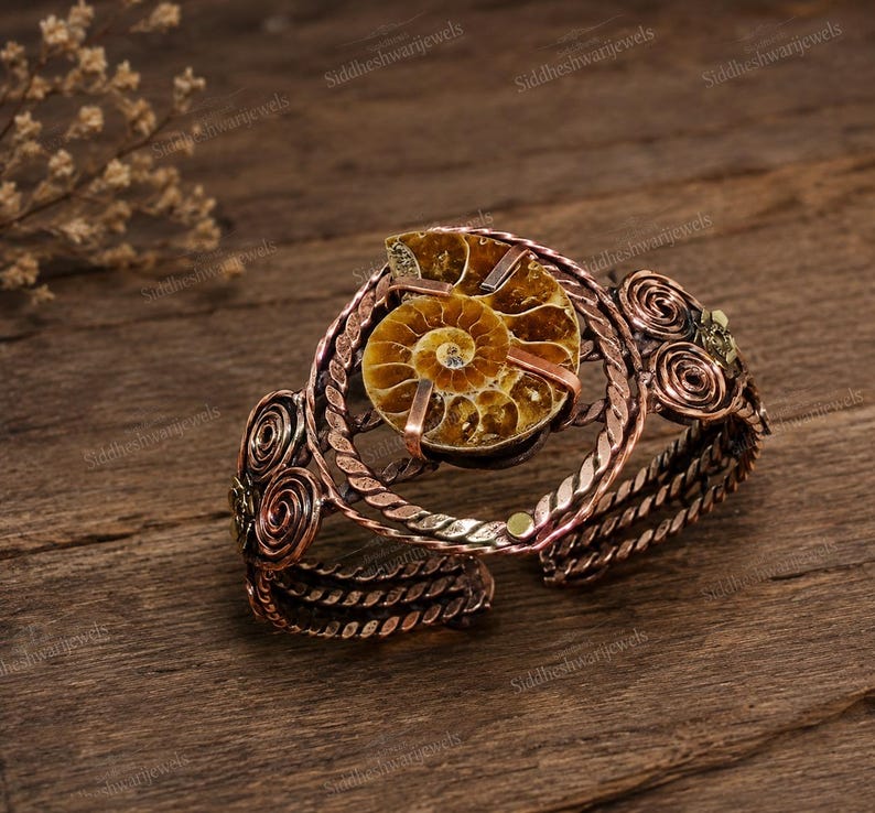 May include: A copper-toned cuff bracelet featuring a central spiral-patterned ammonite fossil held by metal prongs. The band is constructed from twisted wire and decorated with symmetrical wire-wrapped swirls on both sides of the fossil centrepiece.