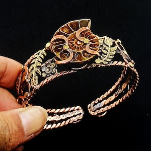 K&ouml;nnte beinhalten: Ein handgefertigtes Kupferarmband mit einem Ammonitenfossil als Mittelst&uuml;ck. Das Armband hat aufwendige Blumen- und Blattdetails in Gold- und Kupfert&ouml;nen. Das offene Design erm&ouml;glicht eine verstellbare Passform. Die Breite des Armbands betr&auml;gt ca. 5 cm.