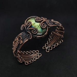 May include: A copper wire-wrapped cuff bracelet featuring a central oval labradorite gemstone with green and blue iridescence. The metalwork consists of intricate coiled wire patterns and a structured band with open loop ends.