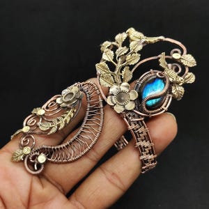 Puede incluir: Anillo artesanal de cobre y latón con una piedra preciosa azul. El anillo presenta detalles florales y de hojas, junto con un diseño de pájaro. La piedra preciosa es de forma ovalada y está engastada en un engaste de alambre.