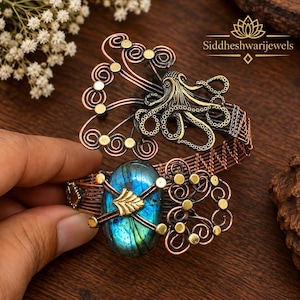May include: A copper wire cuff bracelet featuring an oval labradorite gemstone with a blue iridescent sheen, accented by a gold-toned metal octopus figure, intricate wire swirls, and small circular studs. The text Siddheshwarijewels is visible.