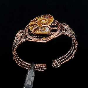 May include: A copper wire cuff bracelet featuring a central polished brown ammonite fossil. The bracelet band is constructed from multiple braided copper wires with intricate spiral wire-wrapped details on the sides, secured by metal clamps.