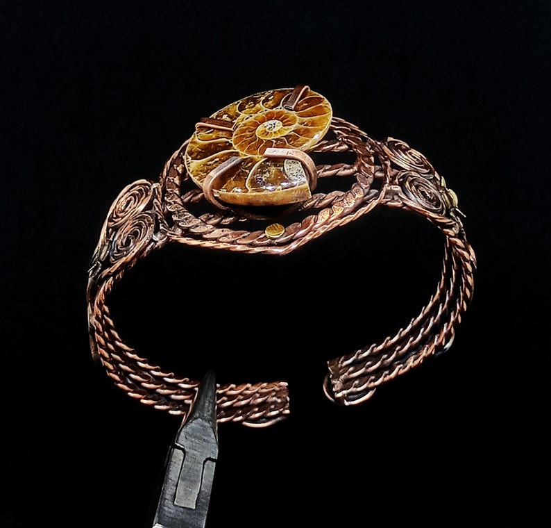 May include: A copper wire cuff bracelet featuring a central polished brown ammonite fossil. The bracelet band is constructed from multiple braided copper wires with intricate spiral wire-wrapped details on the sides, secured by metal clamps.