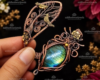 May include: A wire-wrapped cuff bracelet featuring a copper finish with two small gold-toned bird figures perched on a branch design. The bracelet includes a large, oval-shaped iridescent labradorite gemstone and small gold-toned flower accents.