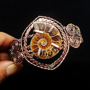 May include: A copper wire-wrapped cuff bracelet featuring a central spiral ammonite fossil. The metalwork includes twisted wire borders and decorative circular coils on the sides, securing the brown and tan fossil with flat metal prongs.