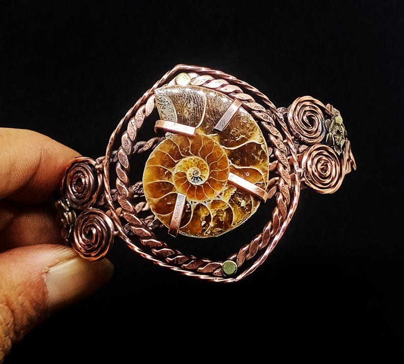May include: A copper wire-wrapped cuff bracelet featuring a central spiral ammonite fossil. The metalwork includes twisted wire borders and decorative circular coils on the sides, securing the brown and tan fossil with flat metal prongs.