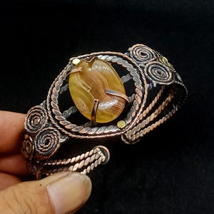 Puede incluir: Brazalete de puño de cobre hecho a mano con una gran piedra preciosa amarilla de forma ovalada. El brazalete tiene un intrincado trabajo de alambre con diseños en espiral y de cuerda, y detalles en metal plateado y dorado. El brazalete es abierto y ajustable.
