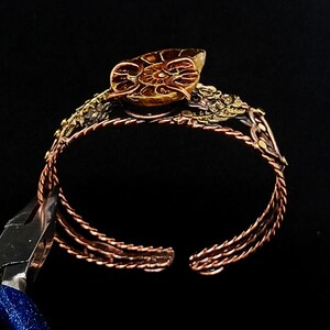 K&ouml;nnte beinhalten: Ein kupferfarbenes Armreif-Armband mit offenem Design. Das Armband verf&uuml;gt &uuml;ber ein detailliertes, fossilienartiges Mittelst&uuml;ck und dekorative Akzente. Das offene Design des Armreifs erm&ouml;glicht ein einfaches Tragen. Das Armband ist vor einem schwarzen Hintergrund platziert.