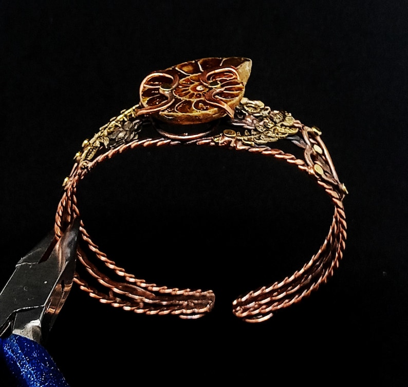 K&ouml;nnte beinhalten: Ein kupferfarbenes Armreif-Armband mit offenem Design. Das Armband verf&uuml;gt &uuml;ber ein detailliertes, fossilienartiges Mittelst&uuml;ck und dekorative Akzente. Das offene Design des Armreifs erm&ouml;glicht ein einfaches Tragen. Das Armband ist vor einem schwarzen Hintergrund platziert.