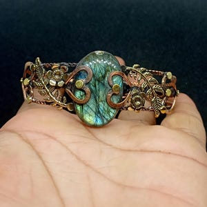 May include: A handcrafted bracelet featuring a large, oval labradorite stone with blue and green iridescence. The bracelet has a copper-colored base with intricate floral and leaf designs in a gold tone, creating a vintage aesthetic.