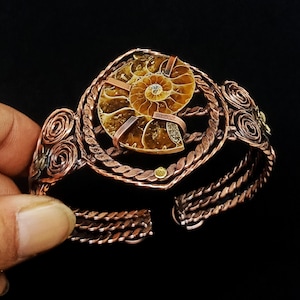 May include: A cuff bracelet crafted from twisted copper wire featuring a central brown ammonite fossil. The design includes intricate wire-wrapped spiral accents on the sides and a braided band structure that secures the fossil in place.
