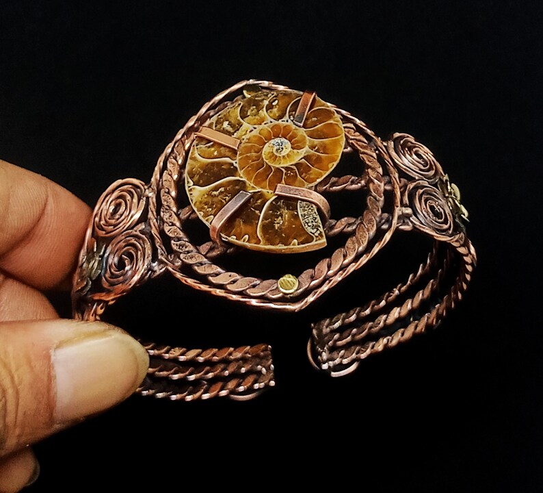 May include: A cuff bracelet crafted from twisted copper wire featuring a central brown ammonite fossil. The design includes intricate wire-wrapped spiral accents on the sides and a braided band structure that secures the fossil in place.
