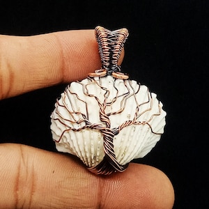 Könnte beinhalten: Handgefertigter Anhänger mit einer weißen Muschel und einem Kupferdraht-Baumdesign. Die Äste und der Stamm des Baumes sind kunstvoll um die Muschel gewebt, mit einer Öse aus Kupferdraht zum Aufhängen. Der Anhänger wird in einer Hand gehalten.