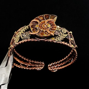 K&ouml;nnte beinhalten: Handgefertigtes Kupferarmband mit einem Ammonitenfossil als Mittelst&uuml;ck. Das Armband hat aufwendige Blumen- und Blattdetails in Gold- und Bronzet&ouml;nen. Das offene Design erm&ouml;glicht ein einfaches Tragen und zeigt die nat&uuml;rlichen Muster des Fossils.