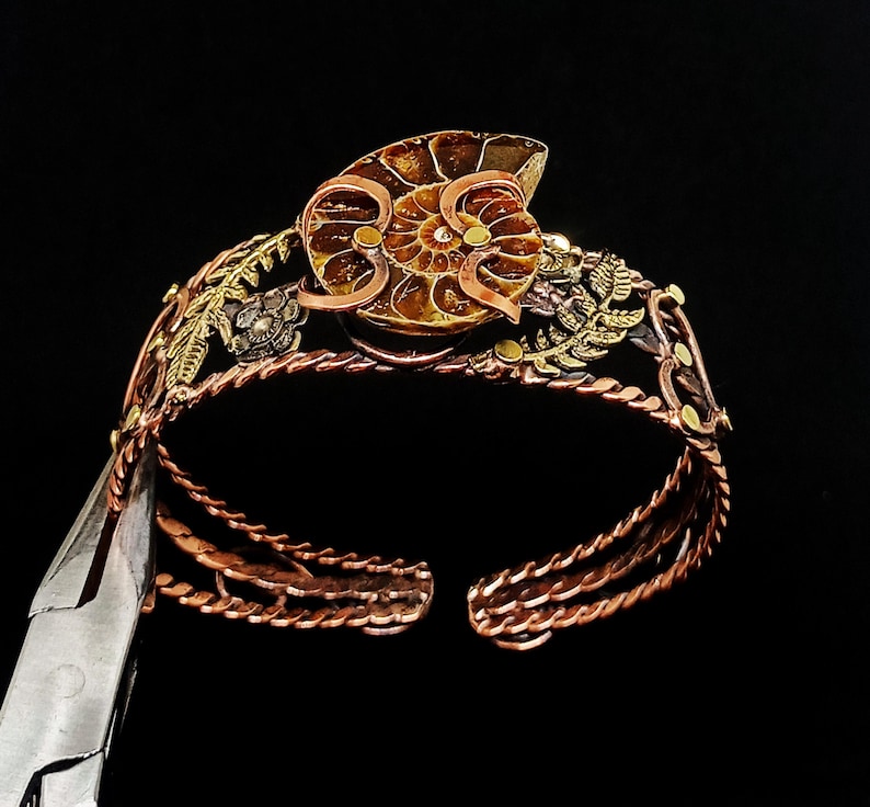 K&ouml;nnte beinhalten: Handgefertigtes Kupferarmband mit einem Ammonitenfossil als Mittelst&uuml;ck. Das Armband hat aufwendige Blumen- und Blattdetails in Gold- und Bronzet&ouml;nen. Das offene Design erm&ouml;glicht ein einfaches Tragen und zeigt die nat&uuml;rlichen Muster des Fossils.