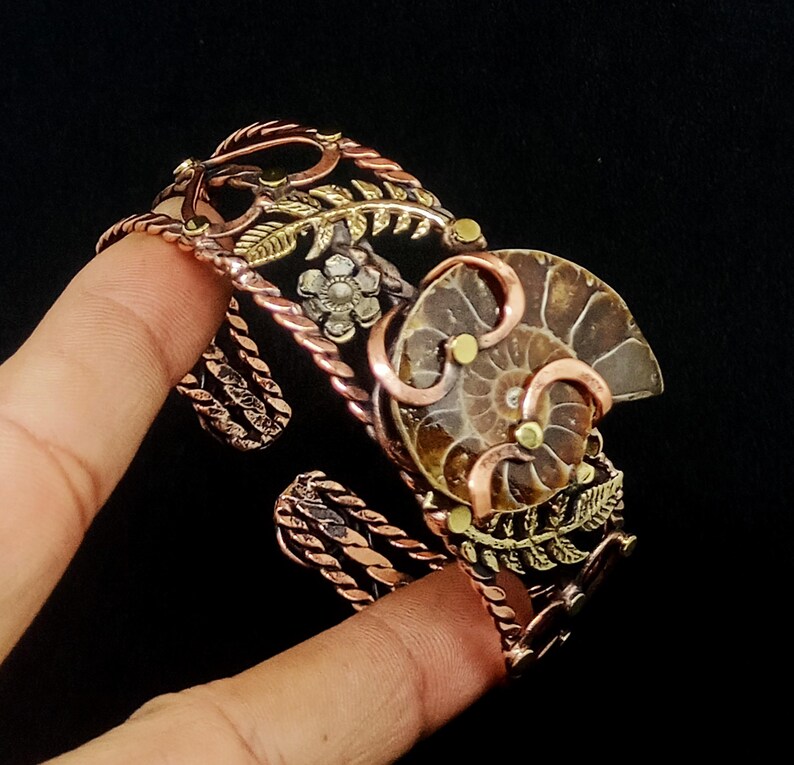 K&ouml;nnte beinhalten: Ein kupferfarbenes Armreif-Armband mit einem Ammonitenfossil-Mittelst&uuml;ck. Das Armband hat aufwendige Blumen- und Blattdetails in Gold- und Silbert&ouml;nen. Das Fossil ist hellbraun, vor schwarzem Hintergrund.