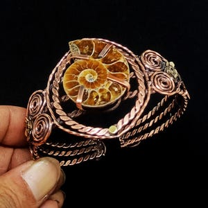 May include: A copper wire cuff bracelet featuring a central circular frame holding a brown spiral ammonite fossil. The bracelet band is constructed from multiple twisted copper wires and decorated with coiled wire accents on both sides.