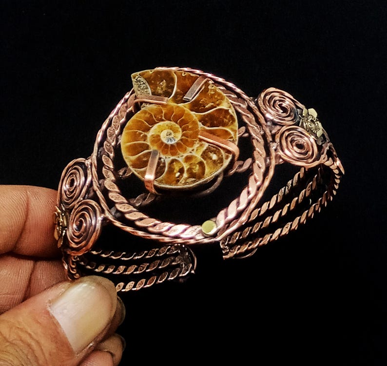 May include: A copper wire cuff bracelet featuring a central circular frame holding a brown spiral ammonite fossil. The bracelet band is constructed from multiple twisted copper wires and decorated with coiled wire accents on both sides.