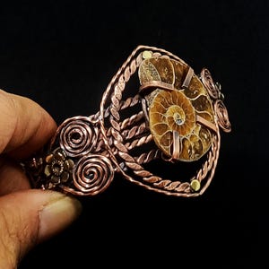 May include: A copper cuff bracelet featuring a central brown ammonite fossil held by metal prongs. The band is constructed from twisted copper wire and decorated with circular wire spirals and small metal flower accents on the sides.