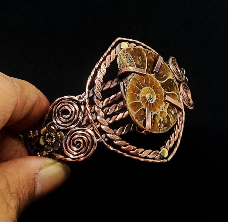 May include: A copper cuff bracelet featuring a central brown ammonite fossil held by metal prongs. The band is constructed from twisted copper wire and decorated with circular wire spirals and small metal flower accents on the sides.