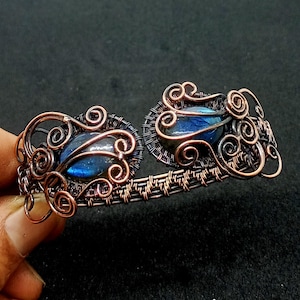 May include: A copper wire wrapped bracelet with two blue labradorite stones set in the center. The bracelet is made with intricate wire work and has a woven design.