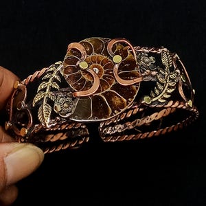 K&ouml;nnte beinhalten: Handgefertigtes Kupferarmband mit einem zentralen Ammonitenfossil-Design. Das Armband weist aufwendige Metallarbeiten mit floralen und Blatt-Akzenten sowie ein gedrehtes, seilartiges Band auf. Die Farbpalette umfasst Kupfer-, Bronze- und Goldt&ouml;ne.