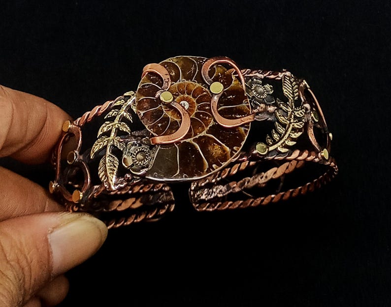 K&ouml;nnte beinhalten: Handgefertigtes Kupferarmband mit einem zentralen Ammonitenfossil-Design. Das Armband weist aufwendige Metallarbeiten mit floralen und Blatt-Akzenten sowie ein gedrehtes, seilartiges Band auf. Die Farbpalette umfasst Kupfer-, Bronze- und Goldt&ouml;ne.