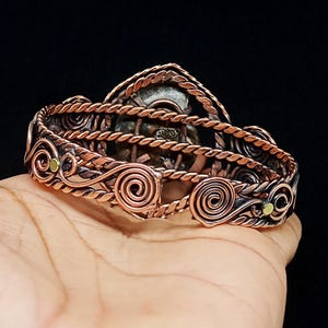 May include: A copper wire-wrapped cuff bracelet featuring a central fossilised ammonite shell. The band is constructed from twisted copper wire and decorative spiral accents, with small brass-coloured circular inlays integrated into the design.
