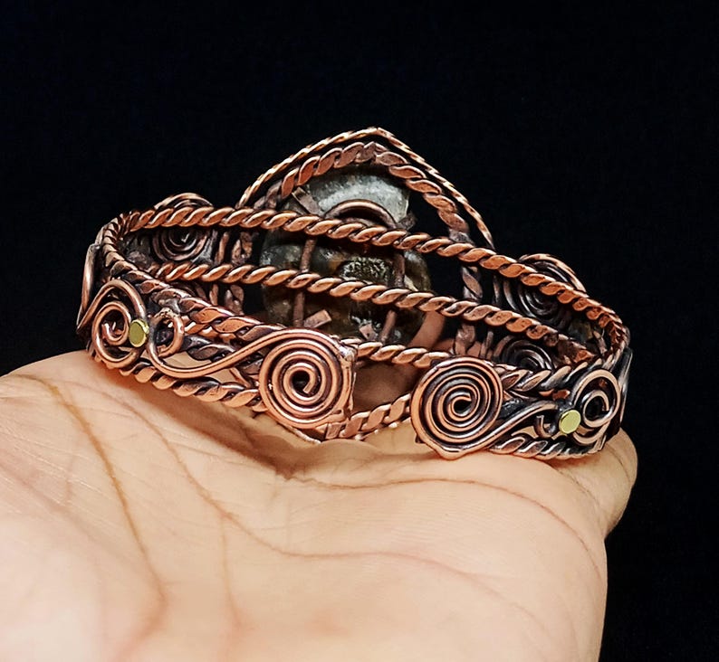 May include: A copper wire-wrapped cuff bracelet featuring a central fossilised ammonite shell. The band is constructed from twisted copper wire and decorative spiral accents, with small brass-coloured circular inlays integrated into the design.