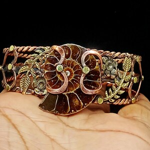 K&ouml;nnte beinhalten: Ein Manschettenarmband aus Kupfer und Messing mit einem gro&szlig;en, braunen Fossilien-Mittelst&uuml;ck. Das Armband hat ein Blumen- und Bl&auml;tterdesign mit einem geflochtenen Seilrand. Kleine gr&uuml;ne Akzente sind sichtbar.